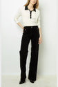 A person wearing a long-sleeve polo blanc with a col noir and buttons, paired with high-waisted black velvet pants featuring a subtle pattern, stands against a plain light background.