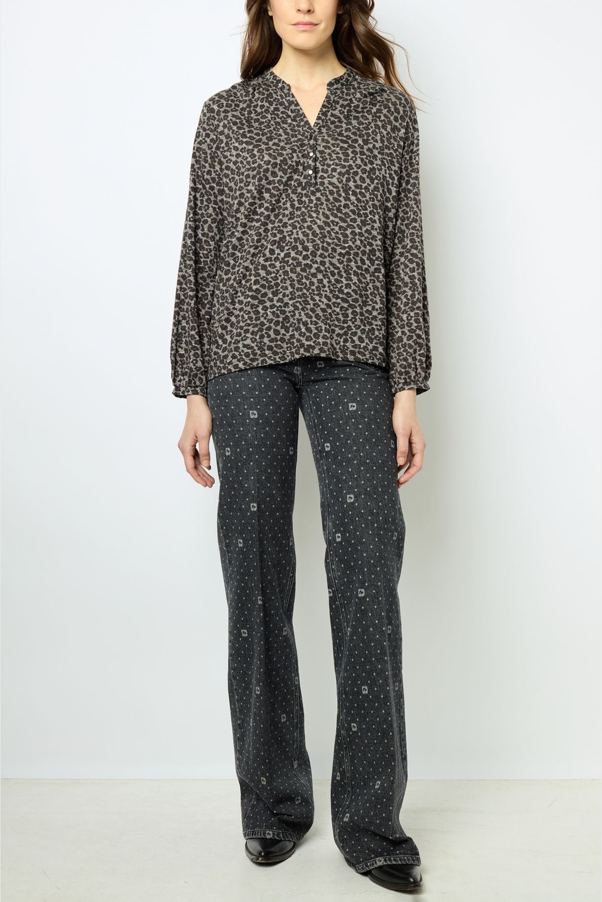 Woman standing against a white background, wearing a long-sleeve, coton mélangé blouse with a subtle pattern and dark, wide-leg pants featuring a small white print, along with black shoes. Her arms are relaxed at her sides.