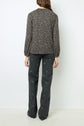 A woman stands facing away, wearing a coton mélangé long-sleeve T-shirt imprimé léopard and dark pants with a subtle geometric pattern. She has long, wavy brown hair and stands against a plain white background.
