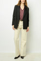 A person stands against a white wall wearing a black blazer, a red patterned blouse, cream-colored wide-leg pants, and black shoes. Their hair is down, and their hands are relaxed at their sides.
