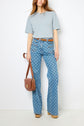 Woman in a light blue t-shirt, patterned high-waisted jeans, tan belt, wooden platform shoes, and carrying a brown shoulder bag, standing against a plain white background.