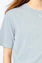 A person with long hair wearing a light blue t-shirt is shown from the shoulders to mid-torso. The logo is visible on the shirt near the neckline. The background is plain white.