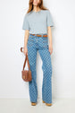 Woman stands against a white wall wearing a light blue T-shirt, patterned blue flared jeans, brown belt, tan shoes, and carrying a brown shoulder bag.