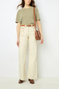 A woman stands against a white wall wearing a khaki t-shirt, cream wide-leg pants, brown belt, brown ankle boots, and a brown crossbody bag.