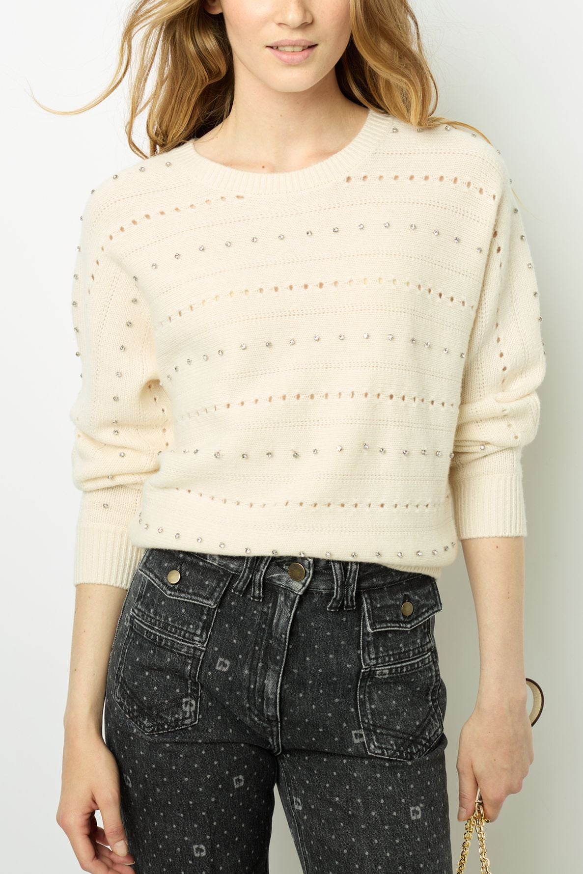 A person wearing a cream-colored knit sweater with small decorative holes and high-waisted black jeans with a subtle dotted pattern. The person is holding a gold chain handbag.