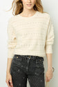 A person wearing a cream-colored knit sweater with small decorative holes and high-waisted black jeans with a subtle dotted pattern. The person is holding a gold chain handbag.