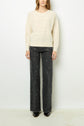 A person stands against a white background wearing a cream sweater with small dots and wide-leg, dark patterned jeans. Their hands rest by their sides, and only their head is partially visible.