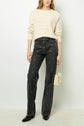 A person wearing a cream sweater, patterned high-waisted black jeans, black pointed shoes, and holding a small beige quilted handbag stands against a plain white wall.
