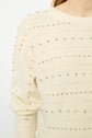A close-up of a cream-colored sweater featuring small silver studs and horizontal open-knit patterns. Only part of the persons upper body and arm is visible.