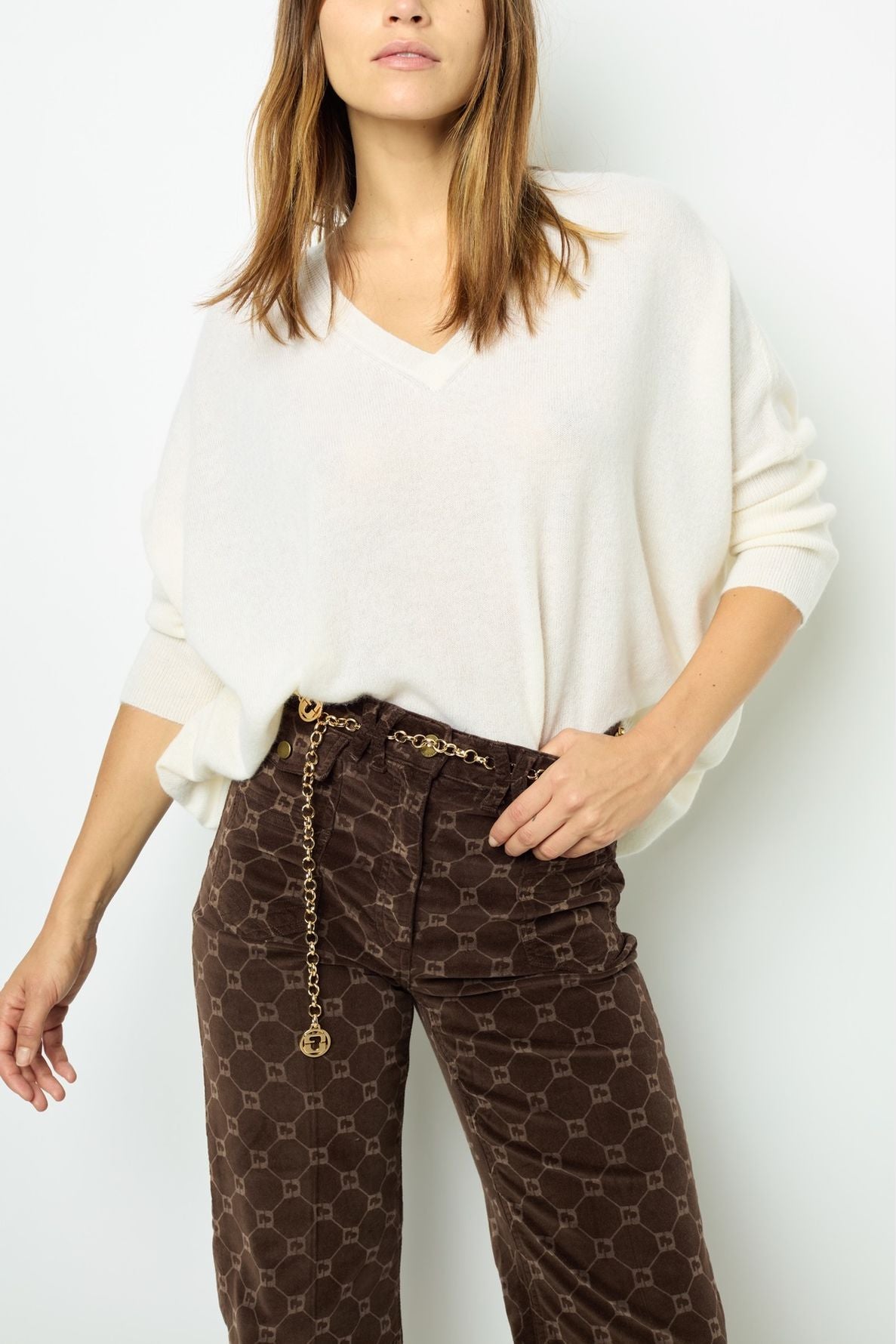 Woman wearing a loose white pull col V en cachemire and brown patterned pants with a gold chain belt, standing against a plain white background.