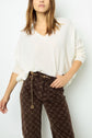 Woman wearing a loose white pull col V en cachemire and brown patterned pants with a gold chain belt, standing against a plain white background.