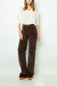 A woman wearing a white pull col V sweater, brown patterned wide-leg pants, a chain belt, and platform shoes stands against a plain white wall. Her face is partially out of frame.