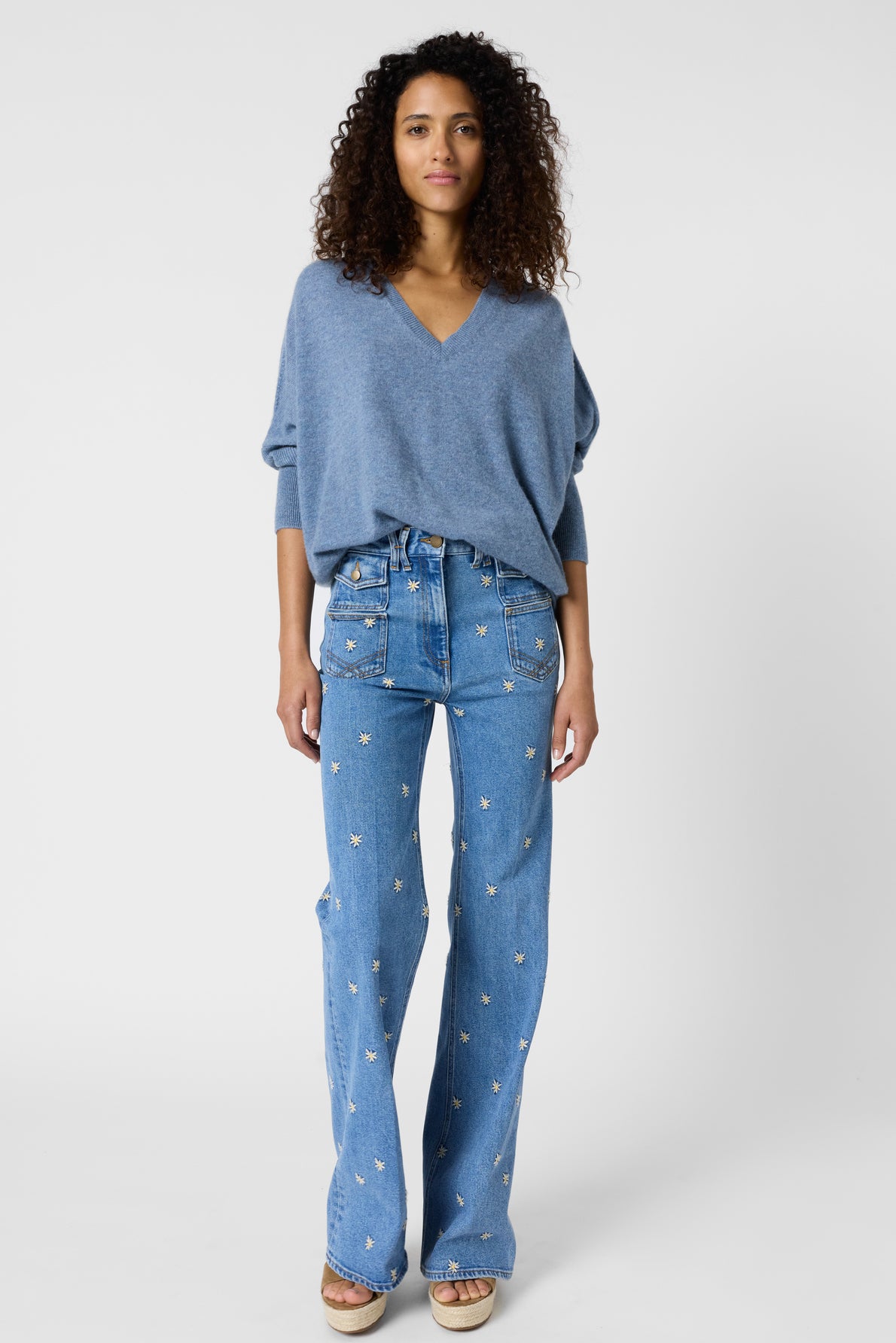 A woman with curly hair wears a loose blue pull col V en cachemire and high-waisted blue jeans with a star pattern, standing on a plain white background.