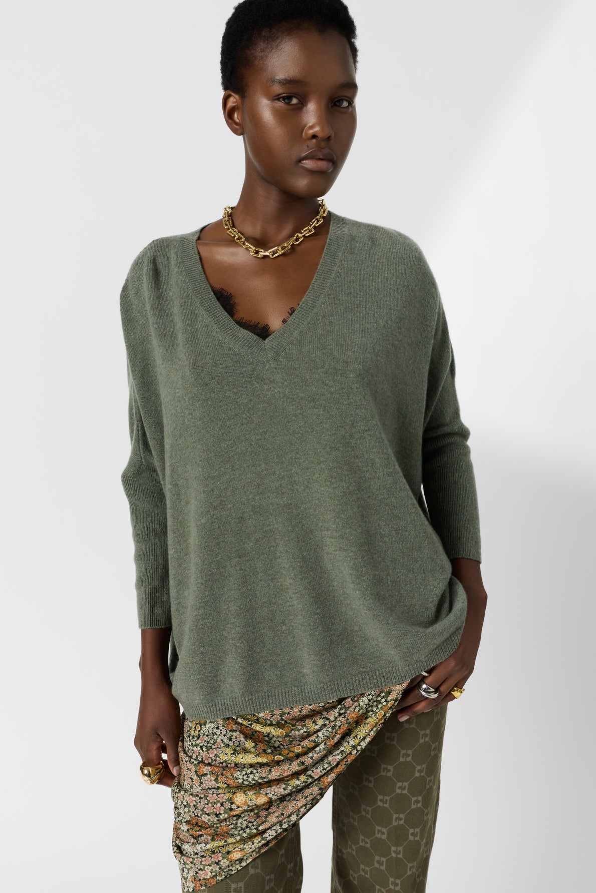 A person wearing a loose green pull oversize col V sweater, gold chain necklace, patterned skirt, and green patterned pants stands against a plain white background.