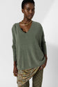 A person wearing a loose green pull oversize col V sweater, gold chain necklace, patterned skirt, and green patterned pants stands against a plain white background.