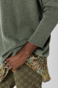 A person wearing a green pull oversize sweater, floral shirt underneath, and patterned olive pants with interlocking Gs, with several rings on their fingers.