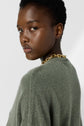 A person with short curly hair wears a green pull oversize sweater and a chunky gold chain necklace, looking over their shoulder toward the camera with a neutral expression against a plain background.