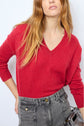 A woman with long, wavy hair wears a pull col V en cachemire rouge profond, tucked into high-waisted gray jeans with multiple pockets, holding a small bag. Only her upper body is visible.