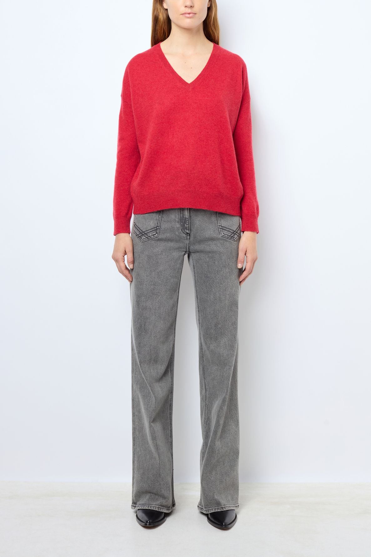 A person wearing a pull col V en cachemire rouge profond, gray wide-leg jeans, and black shoes stands against a plain white background.