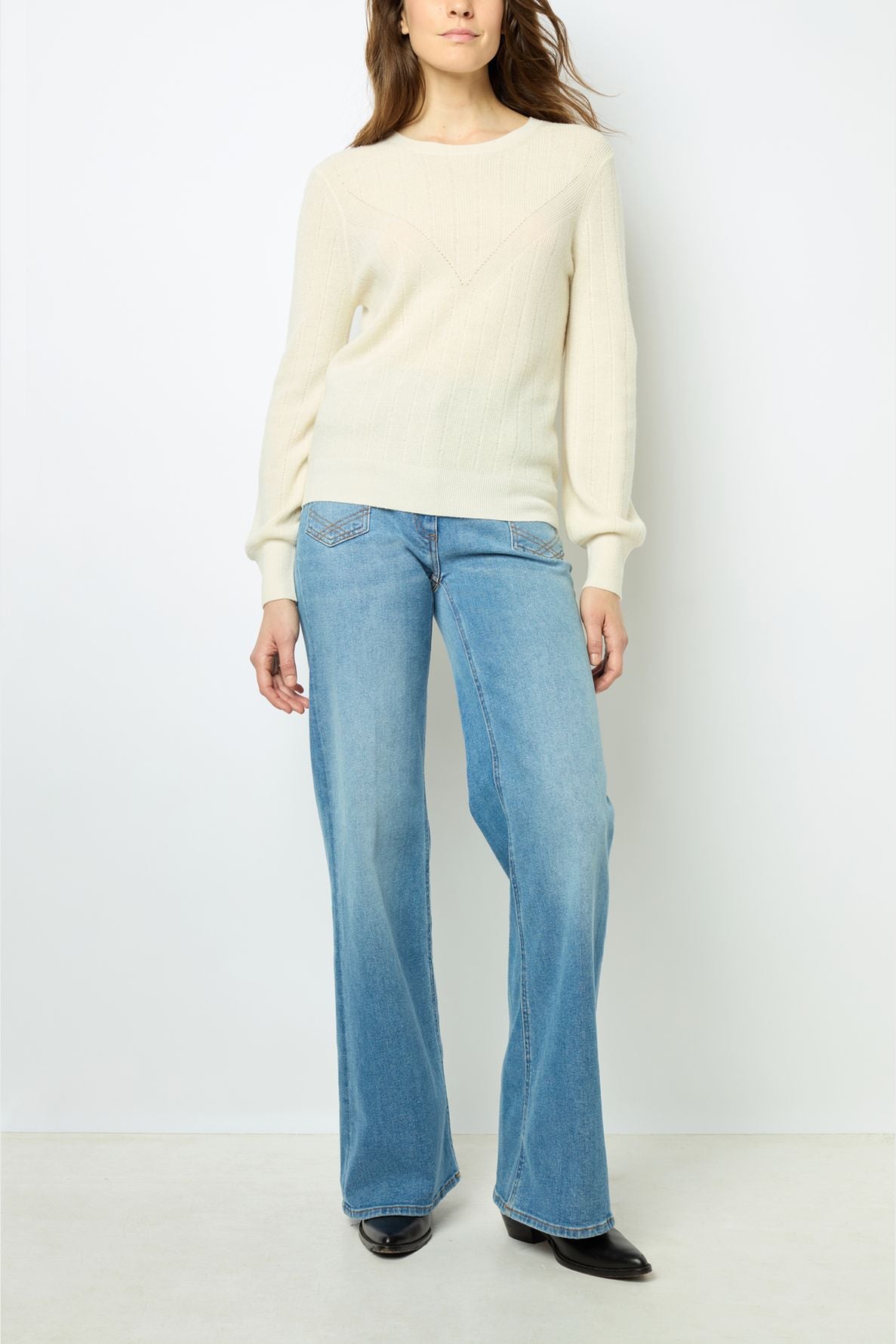 Woman wearing a cream knit sweater and light blue wide-leg jeans stands against a white background, with her hands relaxed by her sides. She is also wearing black pointed-toe shoes.