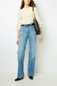 Woman wearing a cream sweater, high-waisted light blue jeans, black shoes, and carrying a black shoulder bag with a gold chain strap, standing against a plain white background.