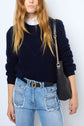 A woman wearing a navy sweater over a white shirt, distressed blue jeans with large front pockets, and a black belt. She carries a black shoulder bag with a gold chain strap.