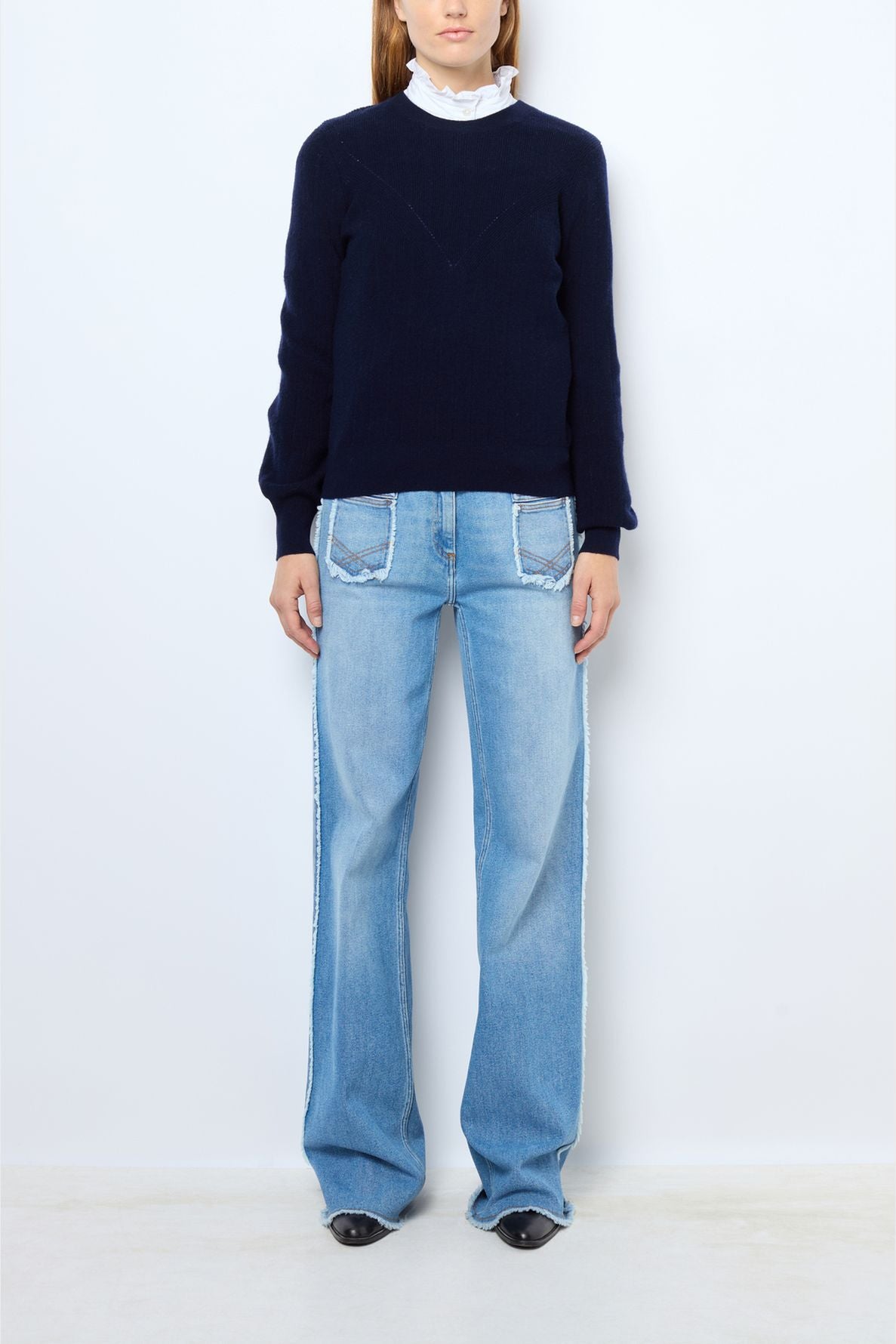 A person stands against a plain white background wearing a navy sweater over a white ruffled shirt and light blue wide-leg jeans with front pockets, paired with black pointed shoes.