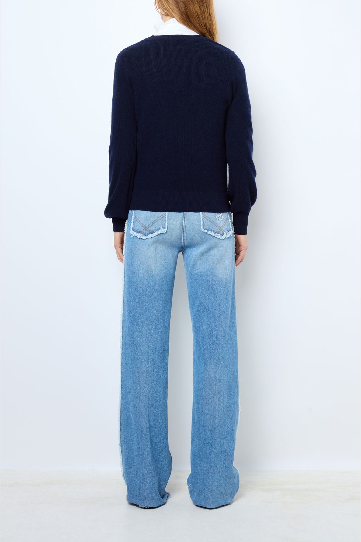 Person standing with back to camera, wearing a navy blue long-sleeve top and light blue wide-leg jeans with decorative back pockets, against a plain white background.