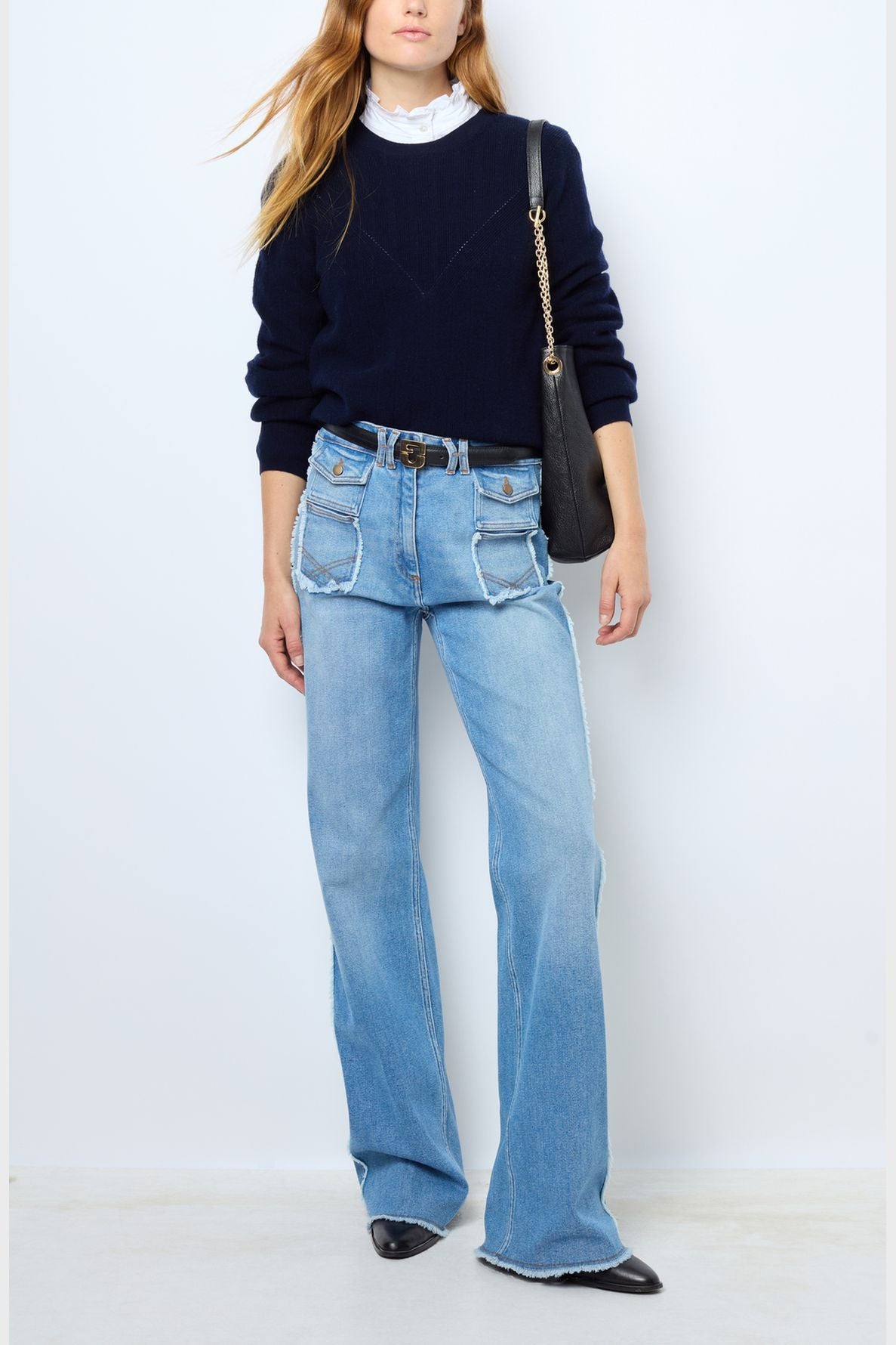 Woman wearing a navy sweater over a white ruffled shirt, light blue high-waisted jeans with large front pockets, black pointed shoes, and carrying a black shoulder bag with a chain strap.