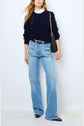 Woman wearing a navy sweater over a white ruffled shirt, light blue high-waisted jeans with large front pockets, black pointed shoes, and carrying a black shoulder bag with a chain strap.