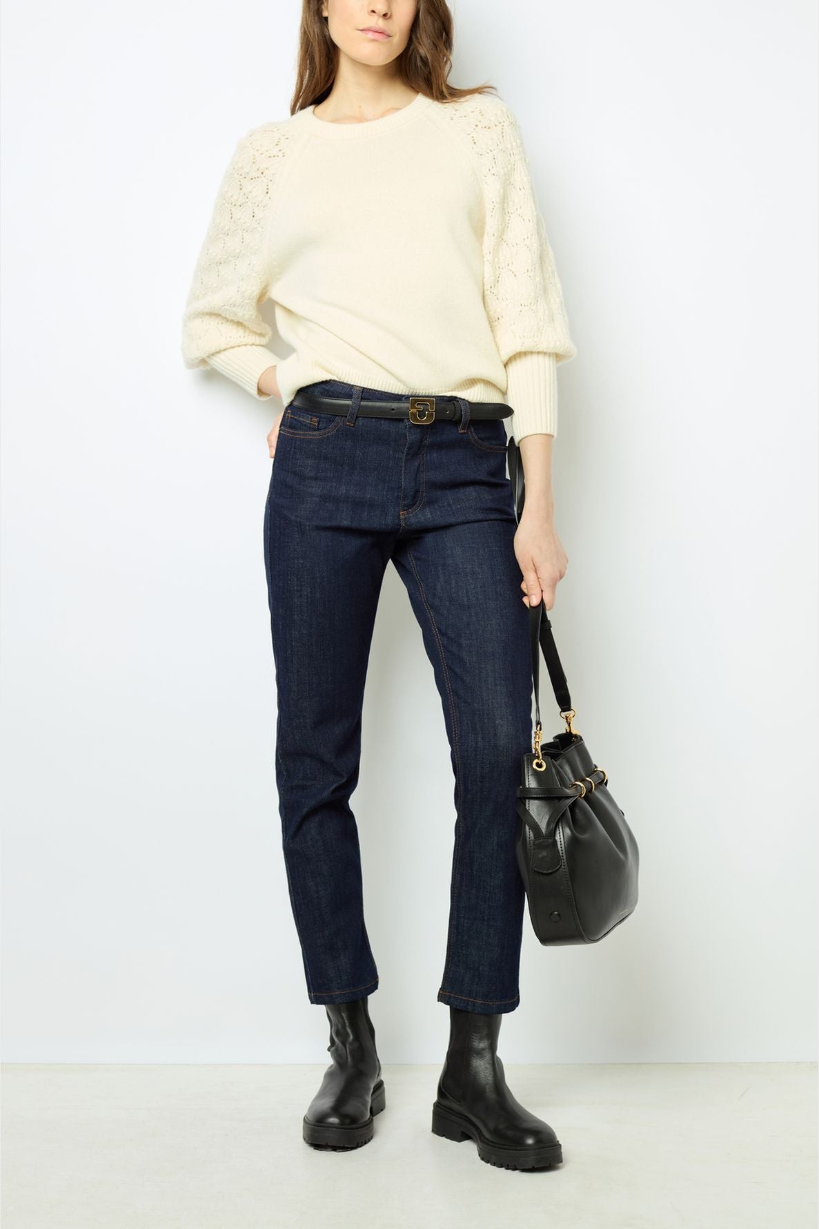Woman wearing a cream sweater, dark blue jeans, and black ankle boots, holding a black handbag, standing against a plain white background.