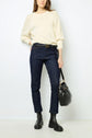 A woman stands against a white wall wearing a cream knit sweater, dark blue jeans, black boots, and holding a black handbag.