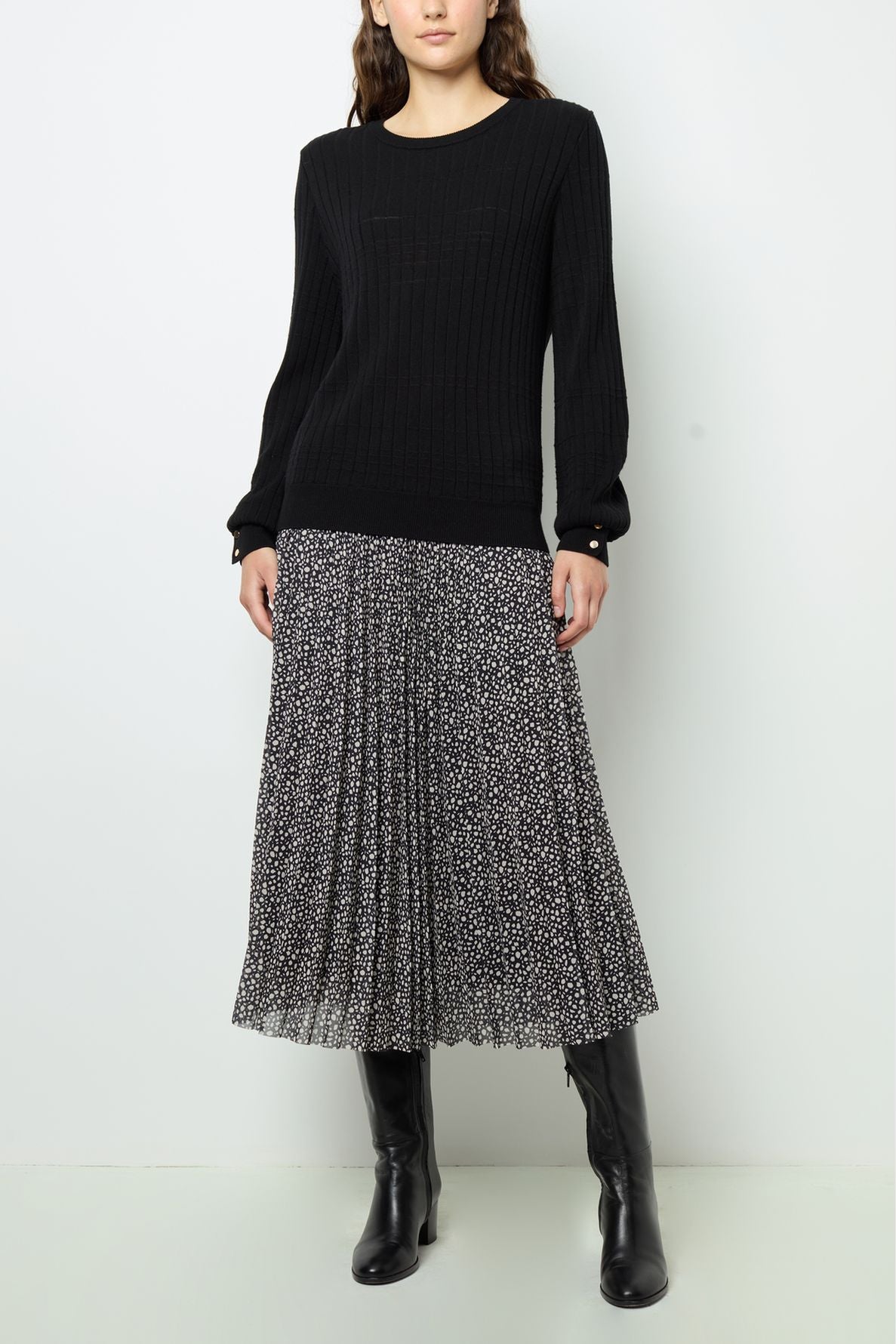A woman wears a black long-sleeve sweater, a black and white pleated midi skirt with a small dot pattern, and knee-high black boots, standing against a plain white wall.