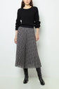 A woman wearing a black ribbed sweater, a black and white pleated midi skirt with a small floral pattern, and knee-high black boots stands against a plain light background.
