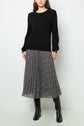 Woman wearing a black ribbed sweater, a black and white pleated midi skirt with a small floral pattern, and knee-high black boots, standing against a plain white background.