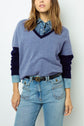 Woman wearing a blue V-neck sweater over a denim shirt, paired with high-waisted jeans and a black belt, standing against a plain light background. Her head is slightly turned and her hair is loose.