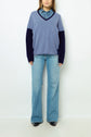 A person wearing a blue V-neck sweater over a denim shirt, light blue flared jeans, and black shoes stands against a plain light background.