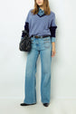 Woman wearing a blue V-neck sweater layered over a denim shirt, light blue wide-leg jeans, black shoes, and carrying a black handbag, standing against a plain white background.