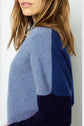 A woman with light brown hair wears a color-blocked sweater in shades of blue, standing sideways against a white background.