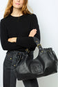 Woman in a black sweater and jeans holding a large black leather handbag with gold hardware and an embossed logo, standing against a plain light background.