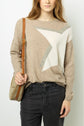 A person wearing a beige sweater with a large white star design, black jeans, and carrying a tan shoulder bag, standing against a plain background.