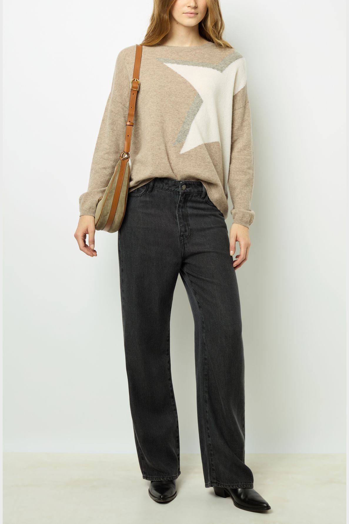 Woman wearing a beige sweater with a large white star design, black wide-leg jeans, black shoes, and a crossbody bag, standing against a plain white background.