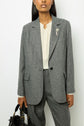 A person wearing a grey blazer à chevrons with a white blouse and a fleur en strass brooch, holding a black handbag, stands against a plain white background.