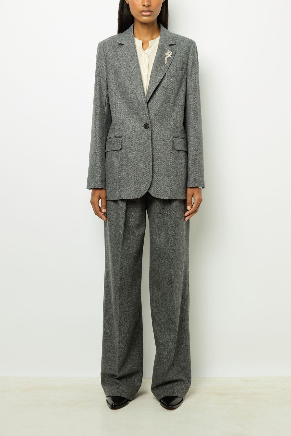 A person stands against a white background wearing a grey blazer à chevrons pant suit, cream blouse, and black shoes. The suit jacket has a fleur en strass brooch on the left lapel. The individual’s face is partially visible.