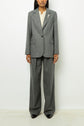A person stands against a white background wearing a grey blazer à chevrons pant suit, cream blouse, and black shoes. The suit jacket has a fleur en strass brooch on the left lapel. The individual’s face is partially visible.