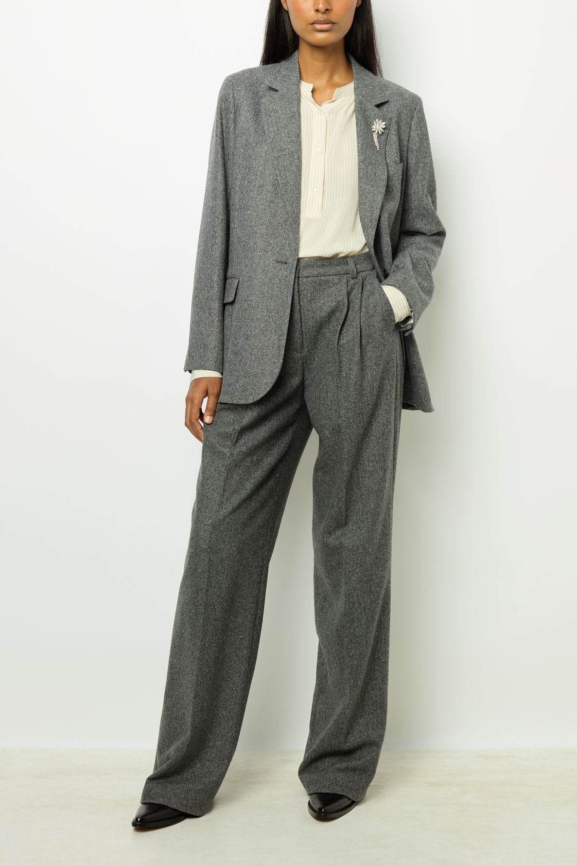 A woman in a gray pantsuit with wide-leg trousers, a cream blouse, and black shoes stands against a plain white background. Her blazer à chevrons features a decorative fleur en strass brooch.