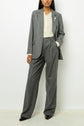 A woman in a gray pantsuit with wide-leg trousers, a cream blouse, and black shoes stands against a plain white background. Her blazer à chevrons features a decorative fleur en strass brooch.
