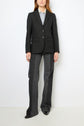 Person wearing a black veste rayée pinstripe blazer, light blue shirt, and dark gray wide-leg trousers, standing against a plain white background. Look channels un esprit tailleur. Face is partially out of frame.