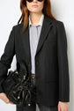 Person wearing a dark blazer imprimé, striped shirt, and sunglasses holds a black leather handbag with gold details. The image is cropped at mid-thigh and set against a plain background.