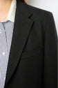 Close-up of a person wearing an esprit tailleur, featuring a dark pinstripe suit jacket over a blue and white striped collared shirt with white buttons. Only part of the face and upper torso is visible.